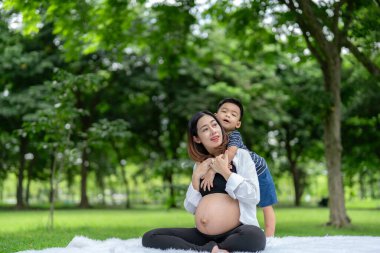 Oğluyla parkta rahatlayan hamile bir kadının portresi. Küçük oğlan annesine arkadan sarılıyor.