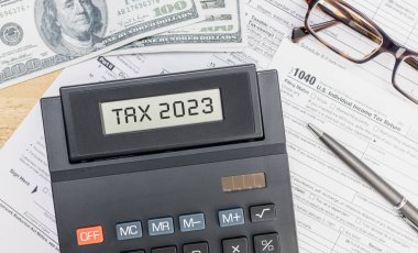 Calculator with TAX 2023 text on screen, 1040 form, dollar banknotes and eyeglasses on desk. Top view