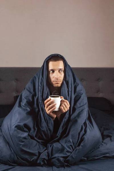 To feel bad. Sick guy with fever, drinking hot healing tea and touching his forehead, lying in bed, empty space. Flu symptom and coronavirus concept