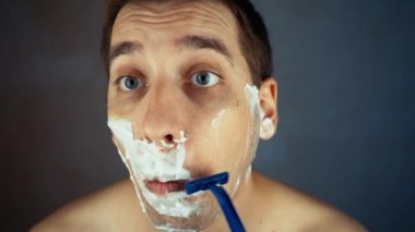 Genç adam banyoda tek kullanımlık tıraş bıçağı kullanıyor, yakın plan. Günlük sabah rutini, hijyenik prosedür. Adam sakal ve bıyık kesiyor.