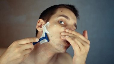 Genç adam banyoda tek kullanımlık tıraş bıçağı kullanıyor, yakın plan. Günlük sabah rutini, hijyenik prosedür. Adam sakal ve bıyık kesiyor.