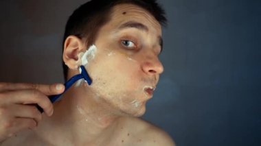Genç adam banyoda tek kullanımlık tıraş bıçağı kullanıyor, yakın plan. Günlük sabah rutini, hijyenik prosedür. Adam sakal ve bıyık kesiyor.