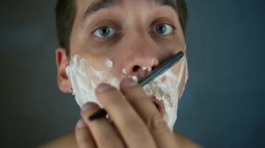 Genç yakışıklı bir adam tehlikeli bir jiletle sakalını tıraş ediyor. Banyoda eski moda bir bıçakla sakalı buduyorum. Yakışıklı maço adam evde sakalını kesti. Yakın çekim.