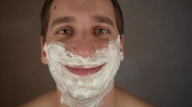Yakışıklı, tıraşsız genç adam nemlendirici sürüyor, yüzüne köpük sürüyor ve kameraya bakıyor. Mutlu bir adamın portresi günlük kişisel bakım yapıyor, içeride güzellik gösterisi yapıyor.
