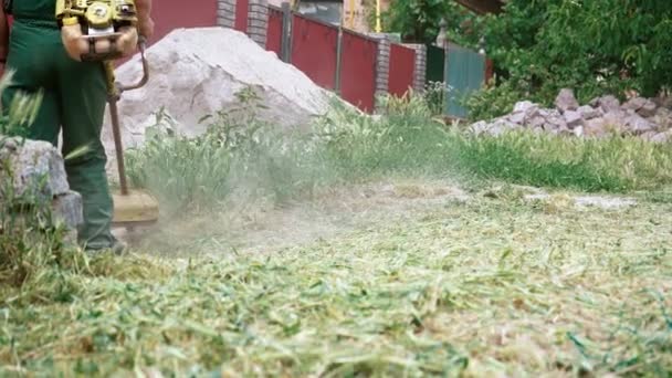 緑の草を刈るトリマーの背景に緑のジーンズの男性の足のクローズアップ 手動のガソリン芝刈り機で草を刈る 庭と芝生のケア — ストック動画