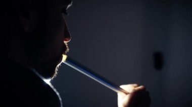 A young man in the backlight smokes a traditional hookah in a dark room, the concept of bad habits. Close-up of a mans face exhaling smoke