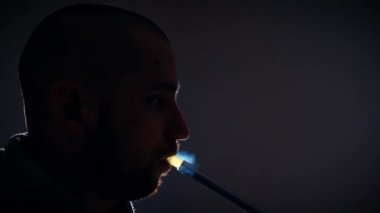 A young man in the backlight smokes a traditional hookah in a dark room, the concept of bad habits. Close-up of a mans face exhaling smoke