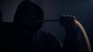 A young man in a hood without a face smokes a traditional hookah in a dark room, the concept of bad habits. Close-up of a mans face exhaling smoke