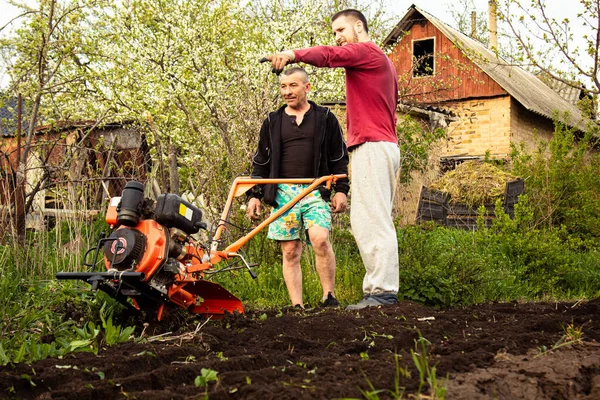 Planting vegetables under the walk-behind tractor. A man with a walk-behind tractor in the garden. Manual work with equipment. An elderly man teaches a young boy to plow the land