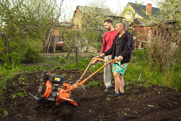 Planting vegetables under the walk-behind tractor. A man with a walk-behind tractor in the garden. Manual work with equipment. An elderly man teaches a young boy to plow the land