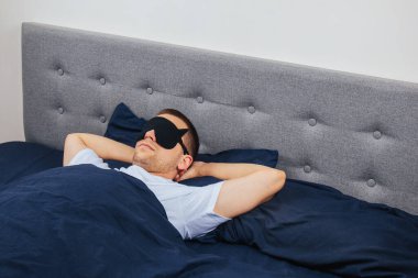 Gri arka planda uyku maskesi takan battaniyeye sarılı bir adam. Evinde, yatağında uyuyor. Dinlenme ve rahatlama kavramı