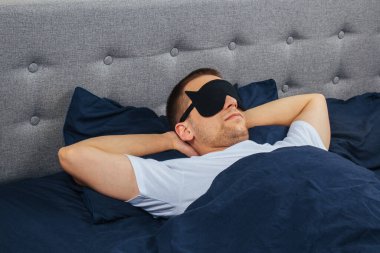 Gri arka planda uyku maskesi takan battaniyeye sarılı bir adam. Evinde, yatağında uyuyor. Dinlenme ve rahatlama kavramı