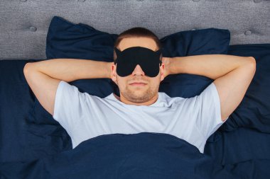Yüksek açılı fotoğraf, uyku maskesi takan genç adam mutlu bir gülümseme, uyku battaniyesi, battaniye, elbiseler, pijamalar.