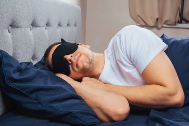 Yatak odasında dinlenen uyku maskesi takan genç bir adamın en iyi görüntüsü. Dinlenmek, işten sonra dinlenmek.