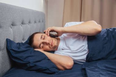 Uyku maskesi takan pijamalı mutlu genç adam. Dinlen, evinde dinlen, otur, battaniye, battaniye altında dön, bir cep telefonunu işaret et, gri bir arka planda izole edilmiş.