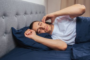 Uyku maskesi takan pijamalı mutlu genç adam. Dinlen, evinde dinlen, otur, battaniye, battaniye altında dön, bir cep telefonunu işaret et, gri bir arka planda izole edilmiş.