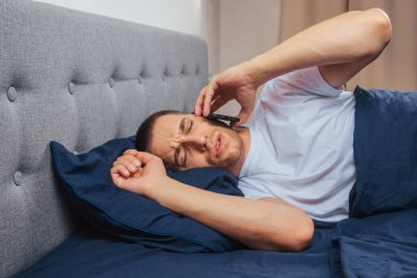 Uyku maskesi takan pijamalı mutlu genç adam. Dinlen, evinde dinlen, otur, battaniye, battaniye altında dön, bir cep telefonunu işaret et, gri bir arka planda izole edilmiş.