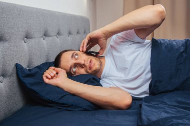 Uyku maskesi takan pijamalı mutlu genç adam. Dinlen, evinde dinlen, otur, battaniye, battaniye altında dön, bir cep telefonunu işaret et, gri bir arka planda izole edilmiş.