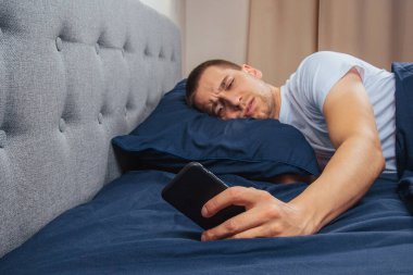 Uyku maskesi takan pijamalı mutlu genç adam. Dinlen, evinde dinlen, otur, battaniye, battaniye altında dön, bir cep telefonunu işaret et, gri bir arka planda izole edilmiş.