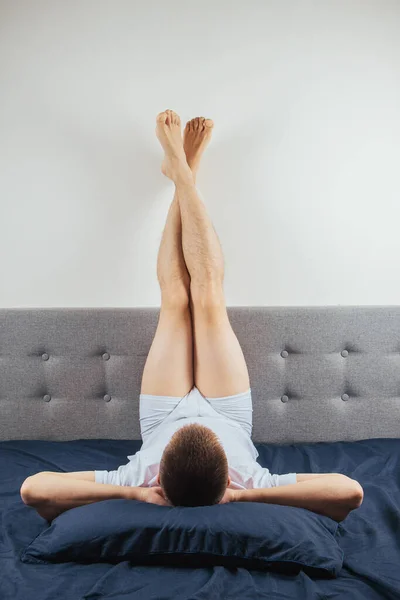 The man lies on the bed with his feet up against the wall. Fooling around and playing, looking at the camera. Resting after a hard day\'s work, relaxation for the legs
