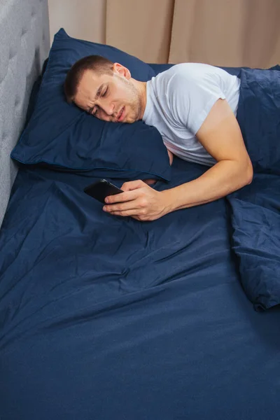 Happy young cheerful man in pajamas wearing a sleep mask. Rest, rest at home, sit, turn around under a blanket, blanket, point a finger at a mobile phone, isolated on a gray background