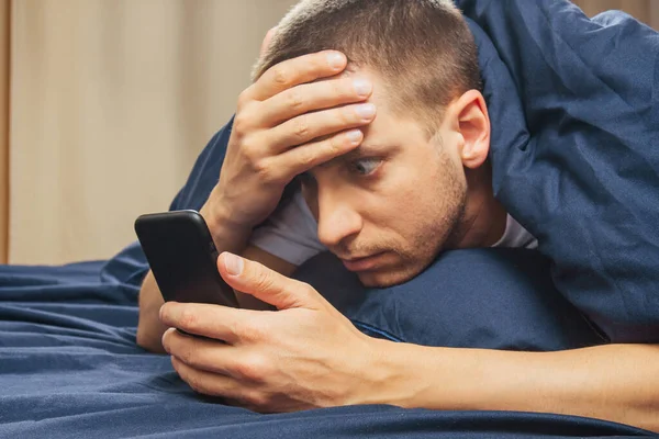 Upset confused young man holding mobile phone, having mobile phone problems or missed call, frustrated angry student reading bad news in message, looking at smartphone, annoyed by spam