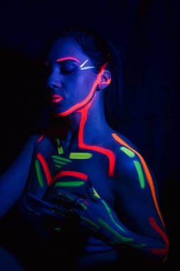 Fashion model woman in neon light, portrait of a beautiful model with fluorescent makeup, body art design in UV, painted face, colorful makeup, on a black background of a girl. Disco dancer in neon light