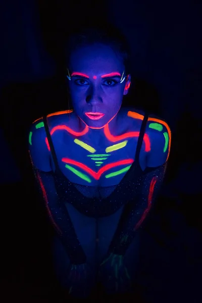 Fashion model woman in neon light, portrait of a beautiful model with fluorescent makeup, body art design in UV, painted face, colorful makeup, on a black background of a girl. Disco dancer in neon light