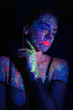 Fashion model woman in neon light, portrait of a beautiful model with fluorescent makeup, body art design in UV, painted face, colorful makeup, on a black background of a girl. Disco dancer in neon light