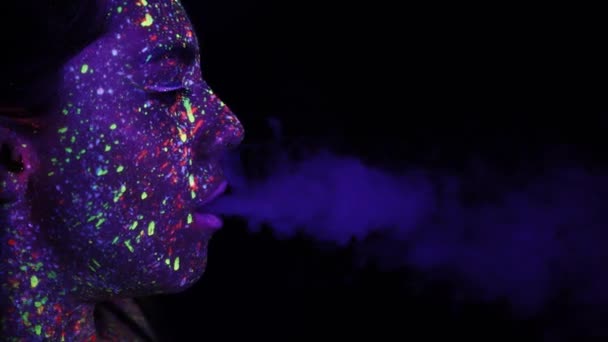 紫外線による化粧をした美少女が煙を描く 口から煙を出す 休日の色のホリー 明るく カラフルで お祝いの着色本 顔の蛍光メイク — ストック動画