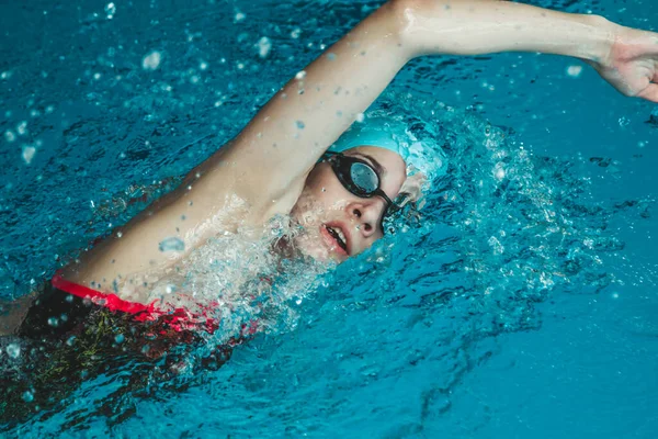 A young swimmer performs a forward crawl technique. The theme of sports and endurance. Side view of a professional female swimmer with goggles at the pool. Close-up. A woman swimmer dives into a pool of water. The girl swims underwater in a large poo