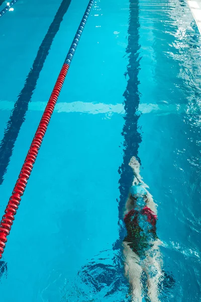 Top view of a young female swimmer training in the pool, swimming on her back underwater. Floats under water and blows air rings