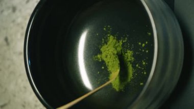 Barista, tahta kaşıkla bir kaseye organik kibrit yeşil çay tozu döküyor. Ahşap bir arka planda siyah bir kase içinde kibrit yeşil çay hazırlama işlemi. Kahverengi bardakta kibrit yeşil çay