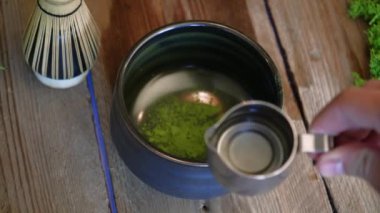 Barista, tahta kaşıkla bir kaseye organik kibrit yeşil çay tozu döküyor. Ahşap bir arka planda siyah bir kase içinde kibrit yeşil çay hazırlama işlemi. Kahverengi bardakta kibrit yeşil çay