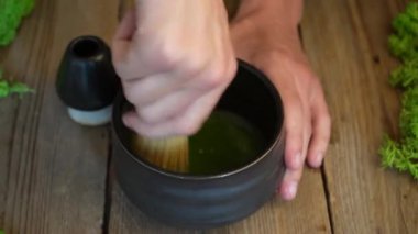 Barista, tahta kaşıkla bir kaseye organik kibrit yeşil çay tozu döküyor. Ahşap bir arka planda siyah bir kase içinde kibrit yeşil çay hazırlama işlemi. Kahverengi bardakta kibrit yeşil çay