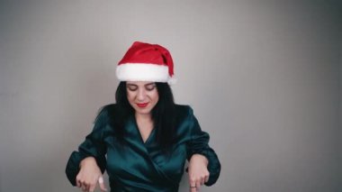 Uzun saçlı, kırmızı Noel Baba şapkası takan ve uzun kollu yeşil gömlekli genç bir kız. Kameraya bakar ve işaret parmaklarını yana çevirir. Noel reklamı. Kış tatili