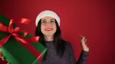 Noel indirimi ve alışveriş. Kırmızı stüdyo arka planında yeşil bir hediye olan Noel Baba şapkalı neşeli bir kadının portresi. Logo için maket. Bağımlı alıcı hepsini satın almak istiyor.