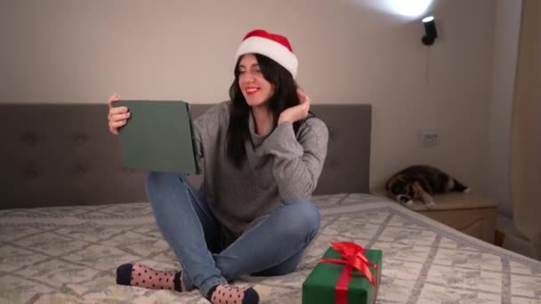 オンラインでのクリスマス挨拶 灰色のセーターと赤いサンタの帽子の女性は オンラインショップにビデオ通話をするためにラップトップを使用しています — ストック動画