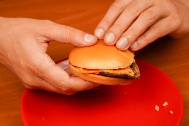 Genç adamın hamburger yerken yakın plan çekimi. Eve teslim. Adam hamburgeri elleriyle tutuyor. Ahşap masada ev yapımı sığır burger tutan eller. Lezzetli fast food hamburgeri yemeye hazır. Sağlıksız gıda konsepti