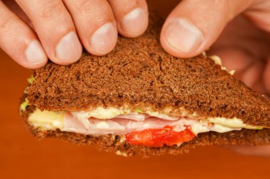 Bir erkeğin yemek pişirirken ve sandviç tutarken yakın plan görüntüsü. Yiyecekler. Lezzetli sandviç. Jambonlu sandviç, pastırmalı, peynirli, marullu, domatesli. Sandviç yiyen adama yakın plan.