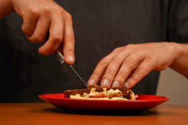 Sandviç yiyen adama yakın çekim. Bir erkeğin yemek pişirirken ve sandviç tutarken yakın plan görüntüsü. Yiyecekler. Lezzetli sandviç. Jambonlu sandviç, pastırmalı, peynirli, marullu, domatesli. Sandviç kesme