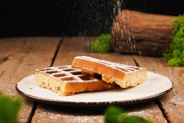 Tabaktaki waffle 'lar koyu arka planda izole edilmiş. Tahta masada Belçika waffle 'ı. Üstü pudra şekerli. Beyaz tabakta Belçika waffle 'ı, ön manzara. Ev yapımı basit hamur işleri.