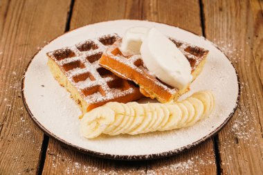 Waffle tabakta. Dondurma sosu. Yemek pişirme süreci. Waffle üzerine muz ekliyorum. Üstü pudra şekerli. Ev yapımı basit hamur işleri. Tahta masada Belçika waffle 'ı. Beyaz tabakta Belçika waffle 'ı