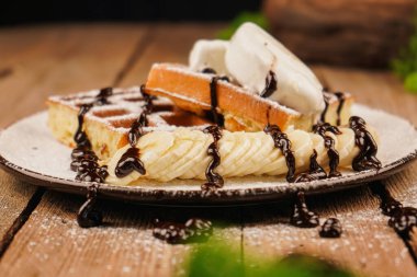 Çay saati için lezzetli bir tatlı. Üstünde muz, dondurma ve çikolata olan Belçika waffleları. Tatlı gözlemeler. Dondurma ile servis edilir. Dondurmalı waffle tabağı ve ahşap masada muz.