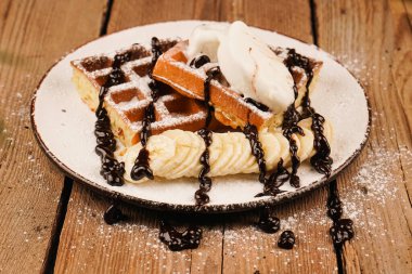 Tatlı gözlemeler. Üstünde muz, dondurma ve çikolata olan Belçika waffleları. Çay saati için lezzetli bir tatlı. Dondurma ile servis edilir. Dondurmalı waffle tabağı ve ahşap masada muz.