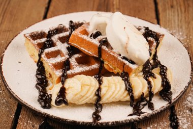 Çay saati için lezzetli bir tatlı. Üstünde muz, dondurma ve çikolata olan Belçika waffleları. Tatlı gözlemeler. Dondurma ile servis edilir. Dondurmalı waffle tabağı ve ahşap masada muz.