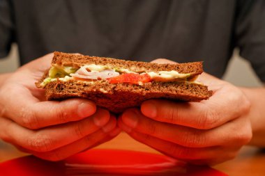 Jambonlu sandviç, pastırmalı, peynirli, marullu, domatesli. Sandviç yiyen adama yakın çekim. Bir erkeğin yemek pişirirken ve sandviç tutarken yakın plan görüntüsü. Yiyecekler. Lezzetli sandviç