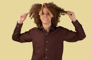 Gülümseyen kıvırcık saçlı sakallı genç adam. Saçlarıyla ilgilenirken kameraya bakıyor. Erken saç kaybı, kepek ya da bit problemleri açık sarı arka planda izole edilmiş..