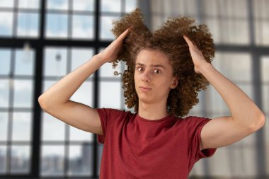 Kıvırcık saçlarından parçalar tutan bir adam. Erkek saç bakım konsepti. Genç Avrupalı adam uzun kıvırcık saçlarını saç fırçasıyla tarıyor. El sallayan saçlarını önemsiyor ve şekillendiriyor. Stüdyonun arka planında duruyorum.