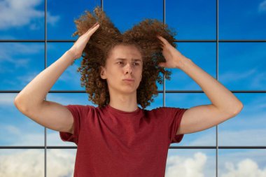 El sallayan saçlarını önemsiyor ve şekillendiriyor. Stüdyonun arka planında dikiliyorum. Kıvırcık saçlarından parçalar tutan bir adam. Erkek saç bakım konsepti. Genç Avrupalı adam uzun kıvırcık saçlarını elleriyle tarıyor.